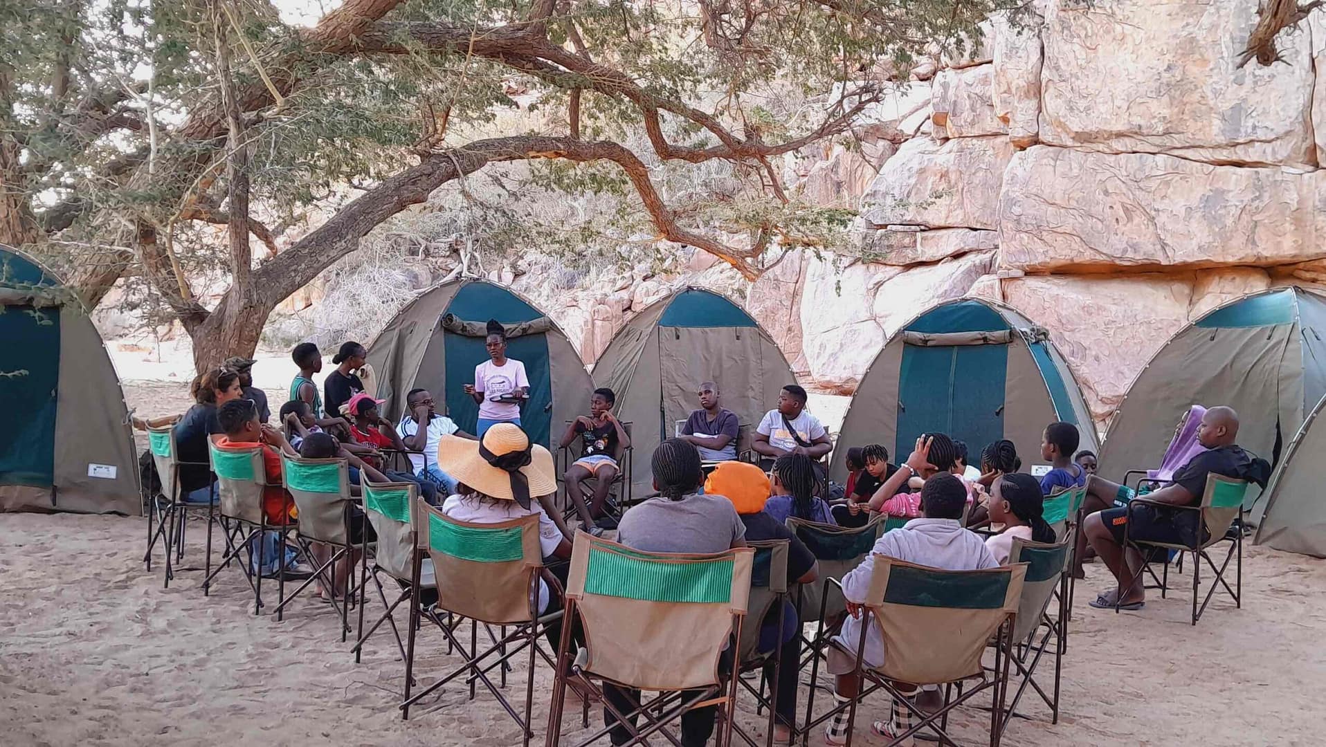 Around 20 people sat at a meeting on camp chairs with tents in the background.