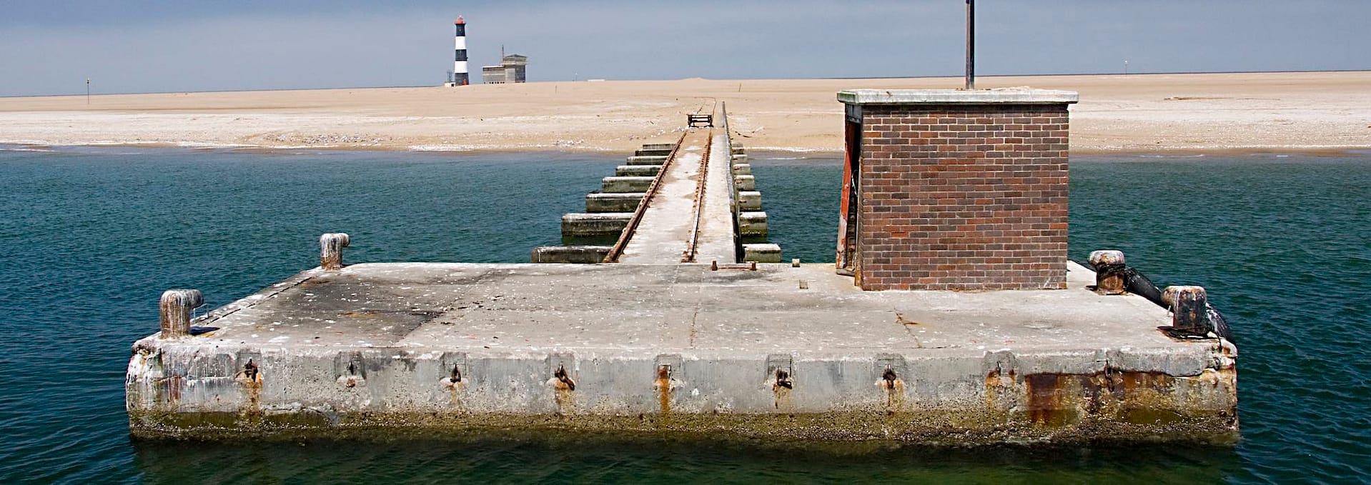 An old concrete pier leading to a sandy beach with a lighthouse in the background.